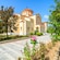 A sunny monastery with a paved pathway surrounded by flowers, trees and topiaries